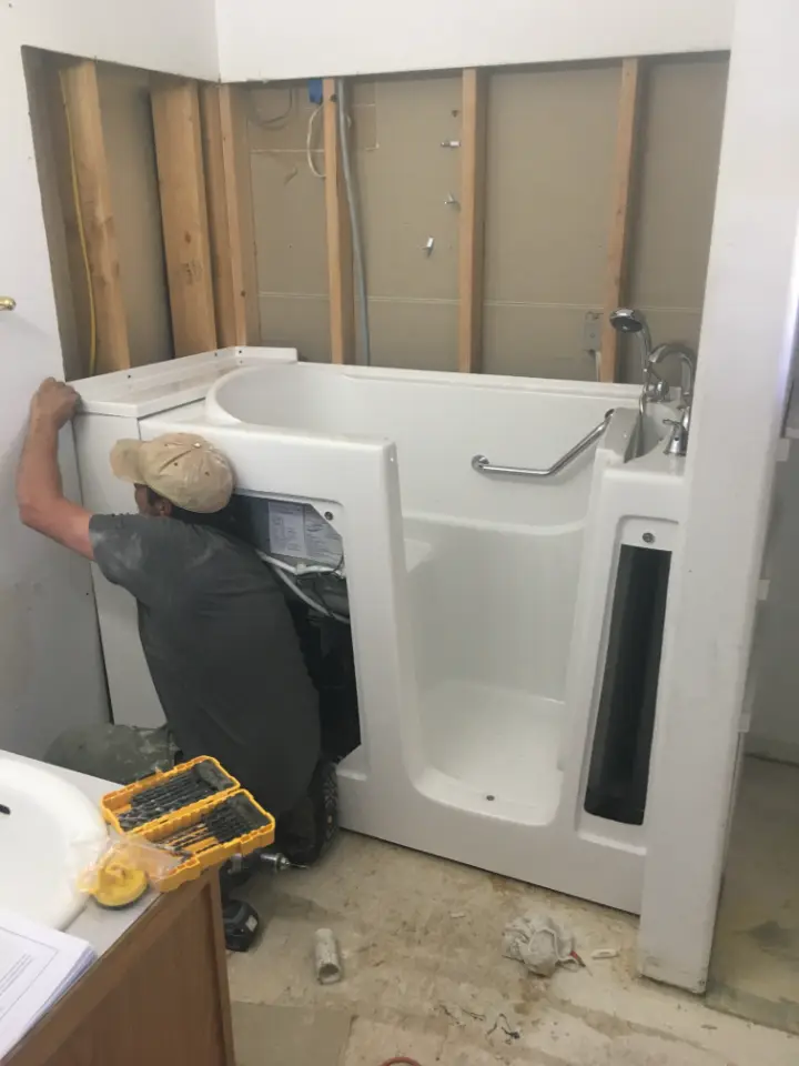 An installer kneels beside a new white walk-in tub set into an alcove with the wall studs exposed, during installation